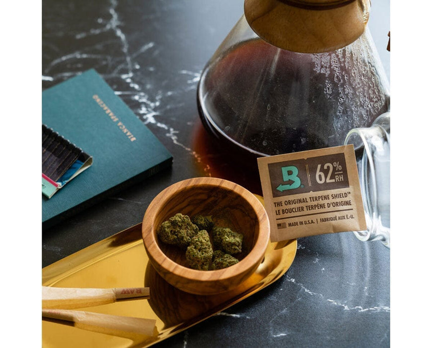 Boveda 62% humidity pack next to buds in wooden bowl and coffee setup, highlighting freshness preservation.