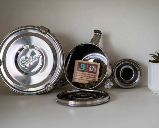 Multiple CVault Grower containers of various sizes shown open and closed, some filled with buds and Boveda packs, arranged against a white background with a small plant nearby.