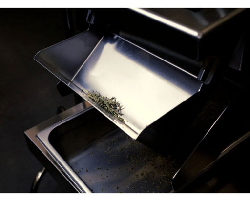 Close-up of GreenBroz Rise-N-Grind output chute dispensing finely ground cannabis trim into a stainless steel tray.