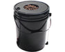 Black 5-gallon hydroponic bucket with a lid containing clay pebbles and a clear water level indicator tube attached to the side.